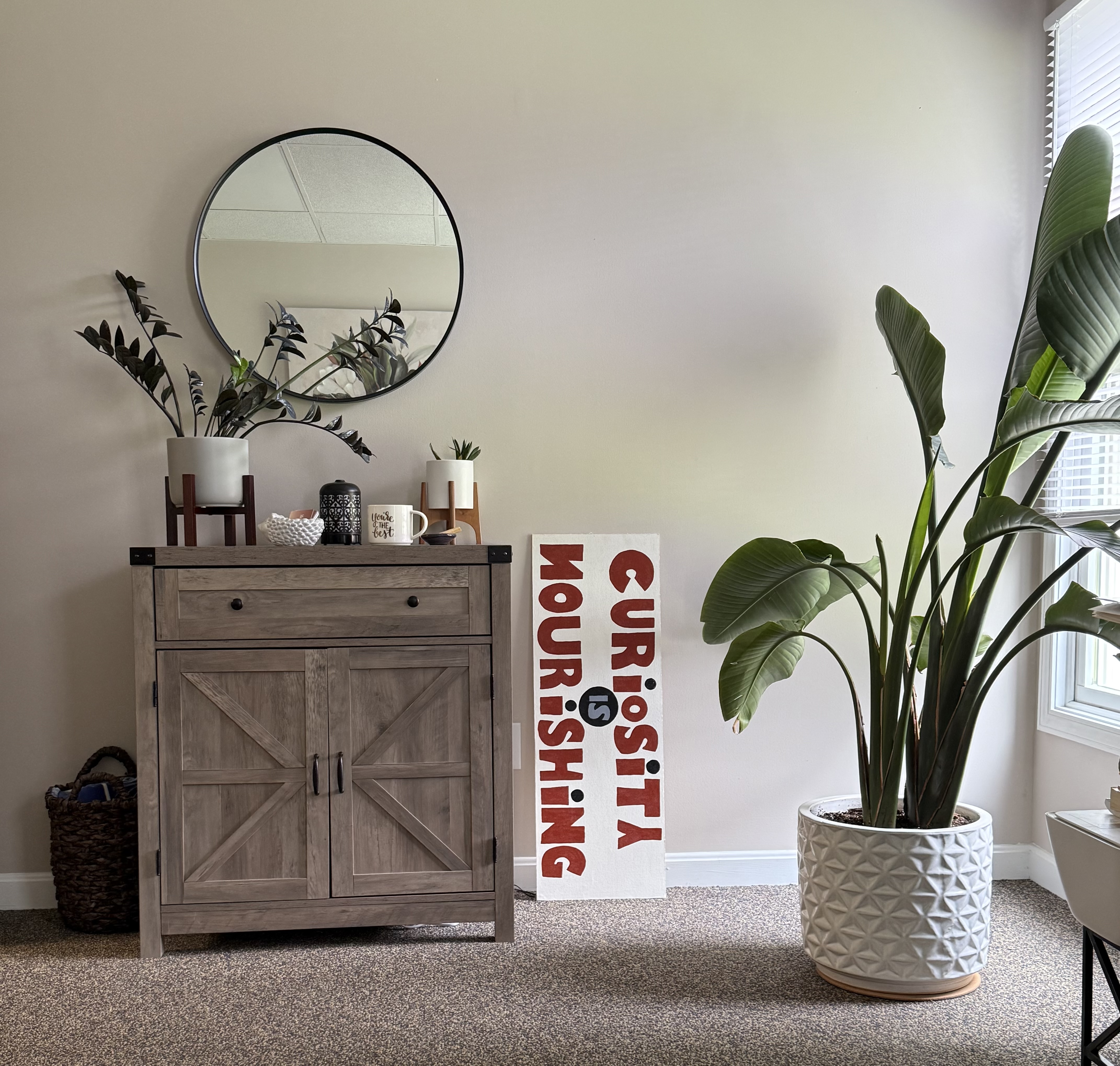 A corner of the therapy office showing plants, a round mirror, rustic sideboard, and a sign reading Curiosity is Nourishing